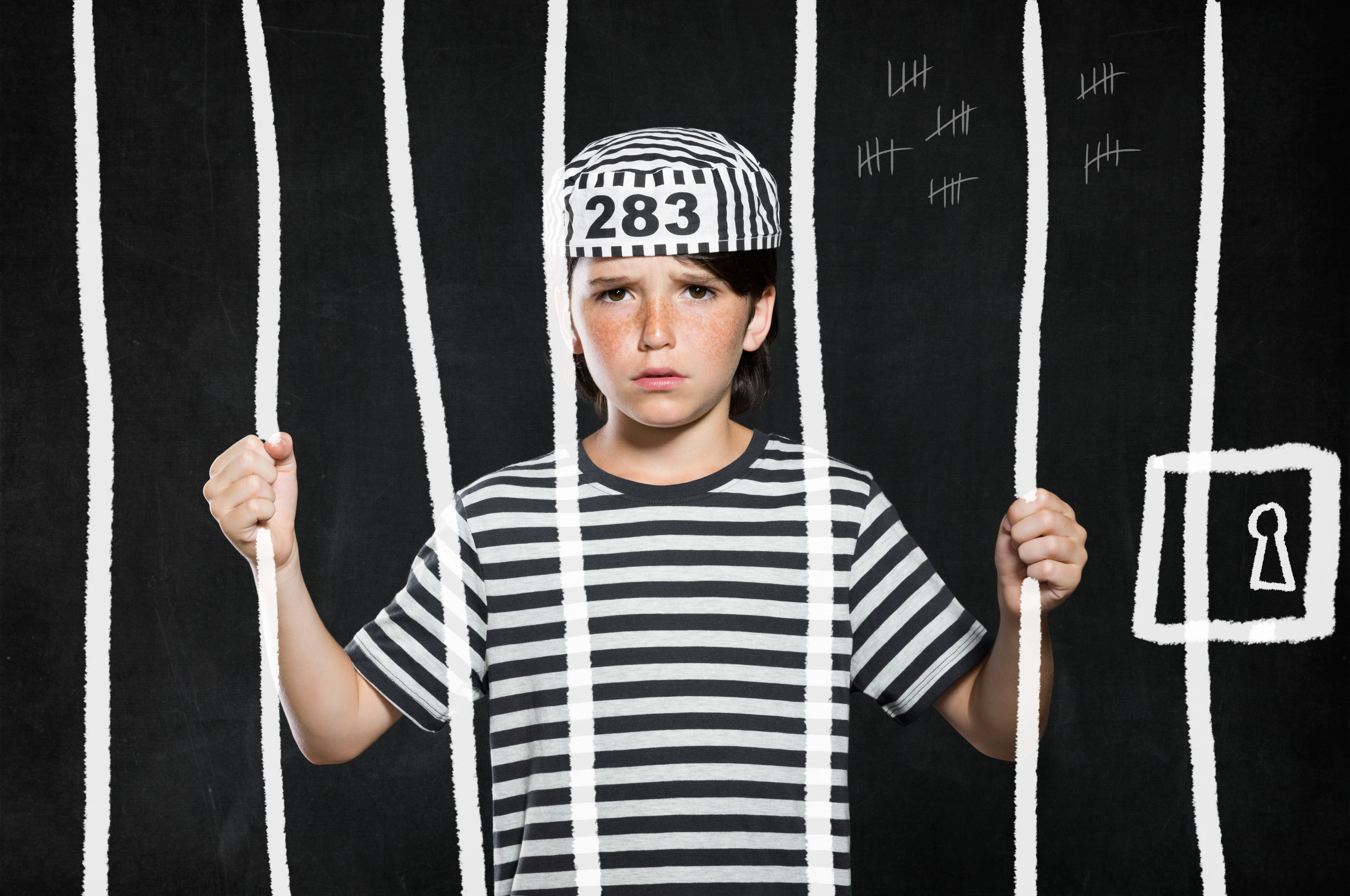 Closeup of sad bad boy wearing jail suit and kept behind bars. Portrait of little prank in jail. Sad male kid wearing striped clothes.